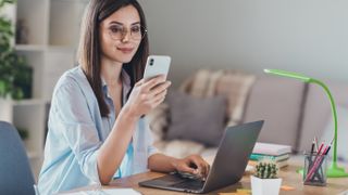 Woman sitting at desk with laptop, holding a phone and looking at it