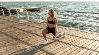 woman in sportswear practicing lunges with dumbbells in hands on the beach at sunrise. Morning workout.