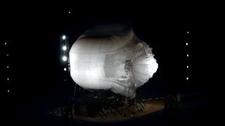 a large white oval shape explodes in a desert at night, surrounded by lights on poles