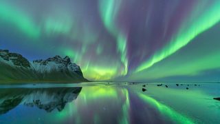 northern lights appear as ribbons of green and purple light streaking across the sky and reflected in the water below. There are some mountains to the left side of the image.