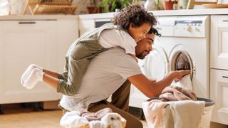 Man and toddler playfully loading a washing machine after stripping the child's bed to remove pee stains from the mattress