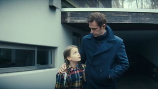 A young girl looking up at her father. He is looking down at her and has his arm wrapped around her shoulder. They are both wearing warm jackets. They are standing next to a house.