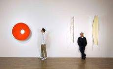 Left: A man looking at a large red circle on the wall with a hole just off of centre. Right: A man leaning back against a wall inbetween two flag-like objects attached to the wall. 