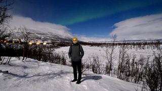 northern lights abisko a lady stands front and center wearing a yellow hat the sky has a distinct gap in the clouds where the northern lights are visible as a river of green through the sky.