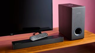 Ultimea Nova S50 soundbar with remote resting on top of it. There is an Amazon Fire TV behind the soundbar, and the Nova S50 subwoofer is positioned to the right of the TV.