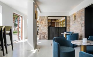 Interior view of another seating area at Bar Roc at Ellerman House featuring white and stone walls, wall lights, blue and green chairs, grey and bronze coloured round tables and a partial view of outside through the open doors. There is also a partial view of the bar area