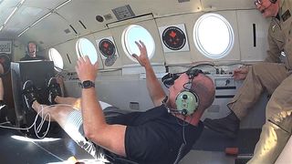 a person lying down in an airplane with their feet on the plate of a flywheel. their hands float, suggesting they are in microgravity during a parabolic flight.