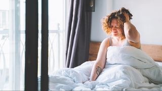 A woman sat up in bed with white sheets next to window with hand on her head as she is waking up with a headache.