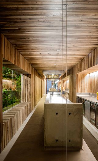 A restaurant kitchen with a long food preparing counter, wooden walls, wooden ceilings, food serving window and ovens and stoves.