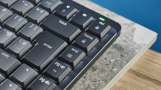 A photo of the Logitech MX Mechanical Mini keyboard in black and gray, on a stone slab and wooden table with a blue background.