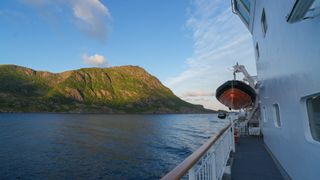 a view from the deck of the hurtigruten ship, you can see a golden mountain with green trees on it to the left and a liftboat on the right.