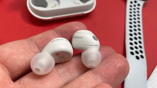 White Sony WF-C700N wireless earbuds in the hand over a red table with their case partially visible in the background.
