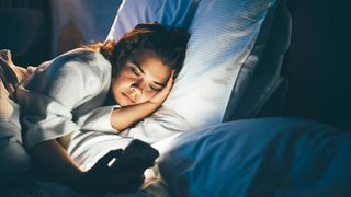 A woman with dark hair lies on her side while scrolling on her phone in bed at night, with blue light shining on her face