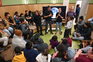 four speakers stand in the center of a round classroom, speaking to a group of adults and children seated around them