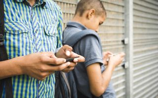 Two teenage boys leaning against a wall texting on smartphones.