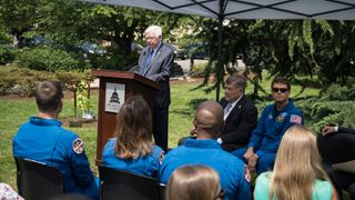 a man in a suit speaks outdoors next to a tiny tree