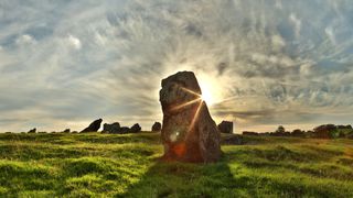 a big stone is in front of the sun's rays. the stone is on grass. Far in the back are other very large stones and trees, and a cloudy sky