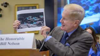 a man in a suit with blue shirt and tie holds up a piece of paper with the image of a cratered moon surface. He has thin, light hair.