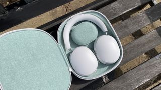 A white pair of Sonos Ace noise-cancelling headphones in their open carry case on a wooden slatted bench.