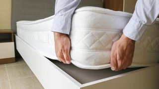 A man lifts one corner of a white mattress to rotate it on a white bed frame