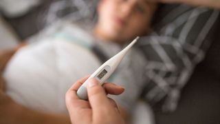 close up of an adult's hand holding a thermometer that reads 100.4. A young boy can be seen in the background under a blanket, as if ill