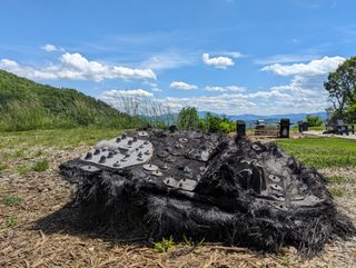 a large black piece of fiberglass covered in metal bolts and plates lies on the ground beside a trail leading into a forest. mountains can be seen rolling in the distance