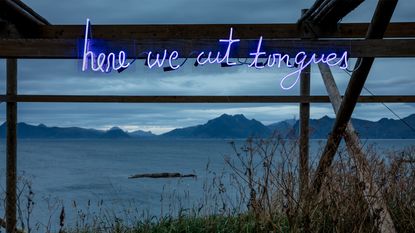 neon letters spelling out ‘here we cut tongues&#039; hung on scaffold in front of lake