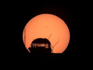 In pitch black, a giant orange orb (the sun) rises behind a domed building under contruction, sihoutted against the glow, with gaps in scaffolding allowing light to pass through the center of the structure. Cranes stand erect and at bent angles on the left and right.