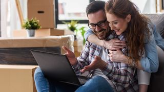 A couple sitting on a couch (a man and a woman), with the man holding a laptop and they're both looking at it