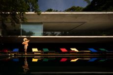 Casa Paraty by Marcio Kogan and studio mk27, shot from the outside pool area looking towards the house with colourful deck chairs and a woman wearing a hat