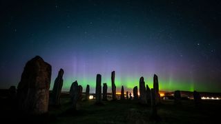 several tall block stones are visible in shadow with the northern lights far in behind. the night sky and stars are visible overhead