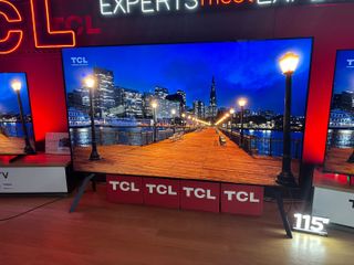 A TCL X955 115 Max TV on display at a trade show. On screen is a lamp post-lined jetty at night with a city skyline in the background.