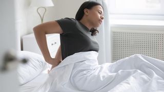 Hard vs soft mattress image shows a woman in a brown t-shirt massages her painful lower back in bed 