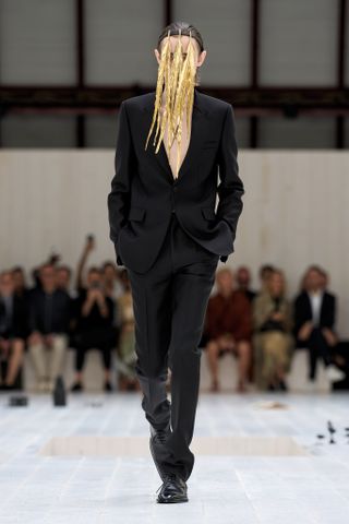 Loewe SS 2025 Menswear runway show featuring model in black suit with gold feathered head piece