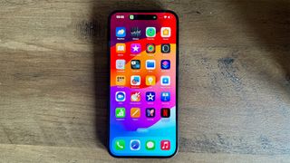 The Apple iPhone 15 Plus smartphone lying on a wooden surface. Its homescreen features a grid of app icons with bands of bright red, orange, purple and blue in the background.