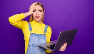 Woman in yellow top clapping hand to forehead while holding laptop.