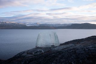 Daytime side view of the Qaammat pavillion, on rocky terrain, water and mountainous terrain in the distance, cloudy sky