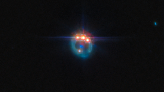 A blurry teal colored gas ring in black space features three bright shining orange orbs at its crown. A blue light dimly shines from the ring's center, and a paler small dot also shines on the right side of the image.