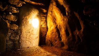 the sun reflects off a large stone inside a barrow. Sunlight falls across the floor and shines against some of the stones nearby. rock walls frame the background