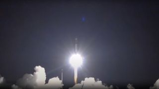 a bright white plume of fire lifts off at night above a cloud of smoke. a white rocket is just barely visible above the flames