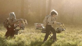 Image from the TV show Star Wars: Skeleton Crew. Two young children walking across a field with their hover bikes. They're both using a walkie talkie.