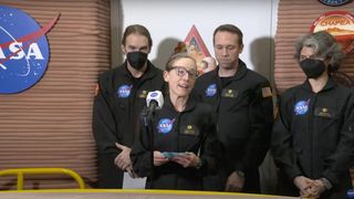 four people in black flight suits stand in front of a microphone, with the nasa logo in the background