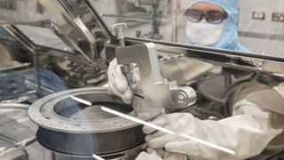 a person in a clean suit and gloves reaches into a glass box to use an L-shaped tool on a round, silver cannister