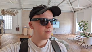 Hamish wearing a black pair of Wayfarer smart glasses from Ray-Ban and Meta. He&#039;s also wearing a hat and a bag in a large modern living room.