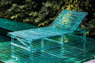 Translucent blue sunbed on a blue tiled pool with green bush in the background