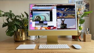 The 2021 Apple iMac (24-inch) in yellow, sat on a wooden desk. The iMac&#039;s desktop is populated with multiple app windows