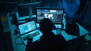 man sat at darkened desk working on laptop and desktop