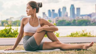 Woman sitting on yoga mat outdoors performing a spinal twist with left leg extended and right leg bent, twisting to the right