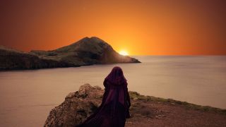 a person in a dark robe stares at a sunrise in the distance over the ocean