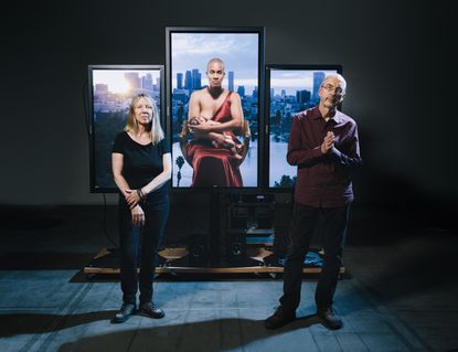 A stand with frames, with focus on the middle frame which shows a woman breastfeeding her baby. A man and a woman are standing in front of the art work